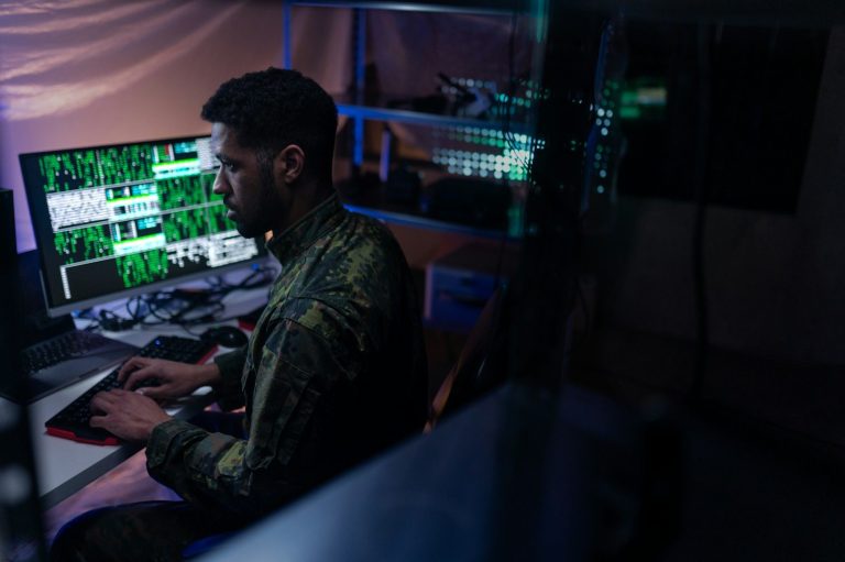 Man wearing military attire working on a computer with multiple green monitors displaying data.