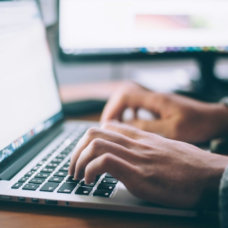Person typing on a laptop keyboard with computer screens in the background.