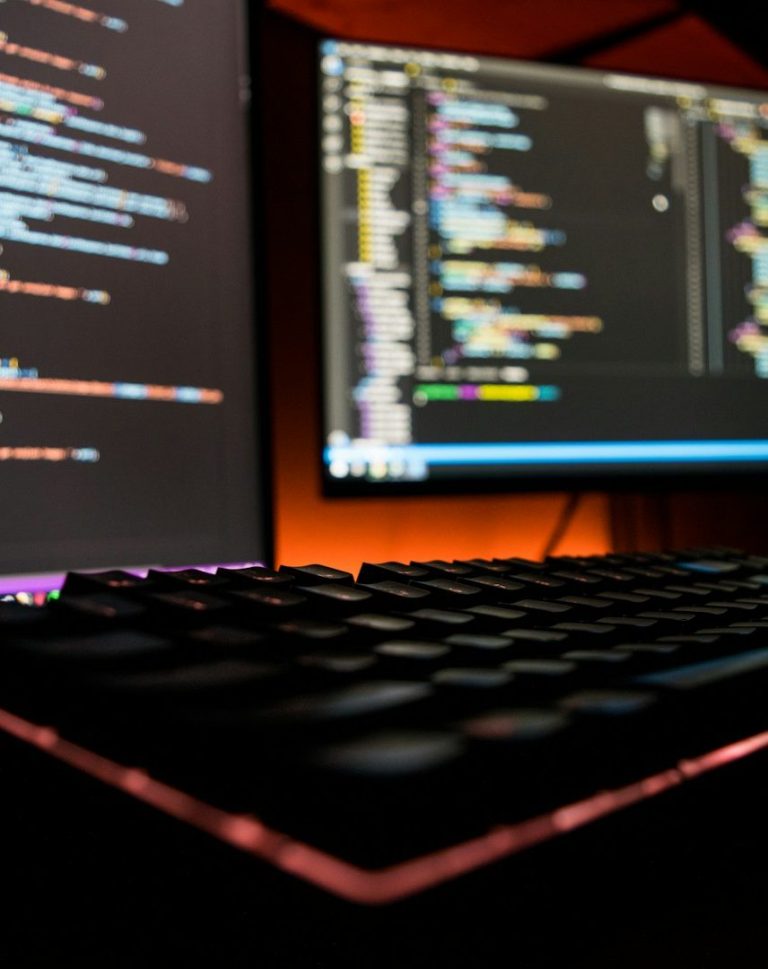 Close-up of a keyboard and monitors displaying coding languages on dark backgrounds.