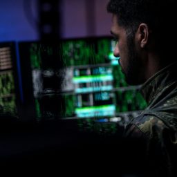 A person in military attire working at a computer with glowing data on multiple screens.