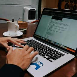 Hands typing on a laptop with a coffee cup and dessert on the table.