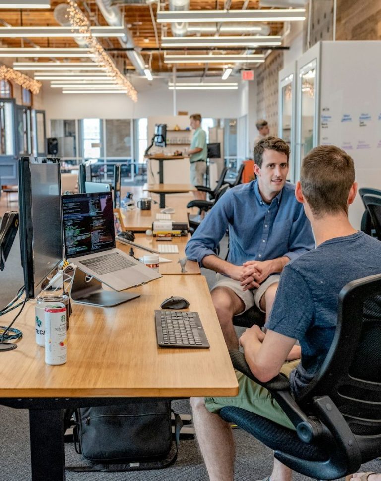 Two men engage in conversation at a modern office workspace with computers.